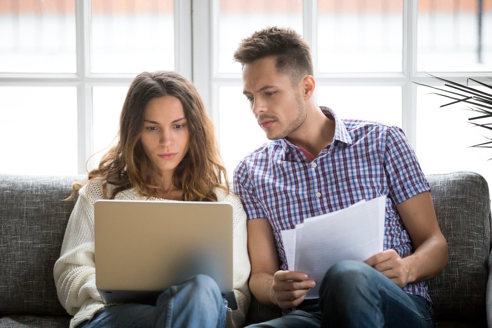 Focused worried couple paying bills online on laptop with documents sitting together on sofa at home, serious confused man and woman planning budget expenses, young family having debt loan problems